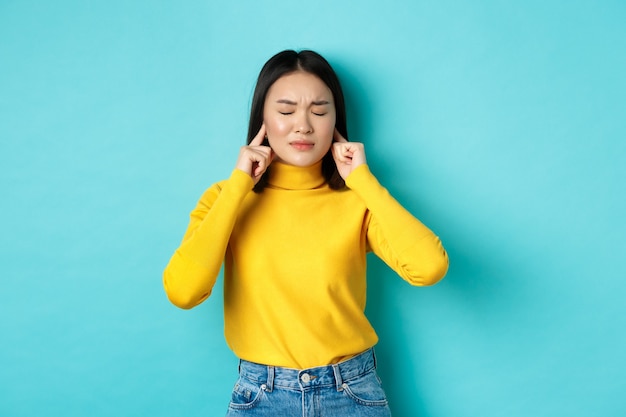 Vermoeide en teleurgestelde jonge aziatische vrouw die niet wil luisteren, oren sluit met vingers en ogen dicht, staande in ontkenning over blauwe achtergrond
