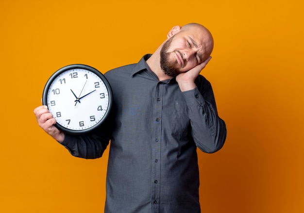 Vermoeide jonge kale callcentermens die klok houdt en slaapgebaar doet dat op oranje muur wordt geïsoleerd