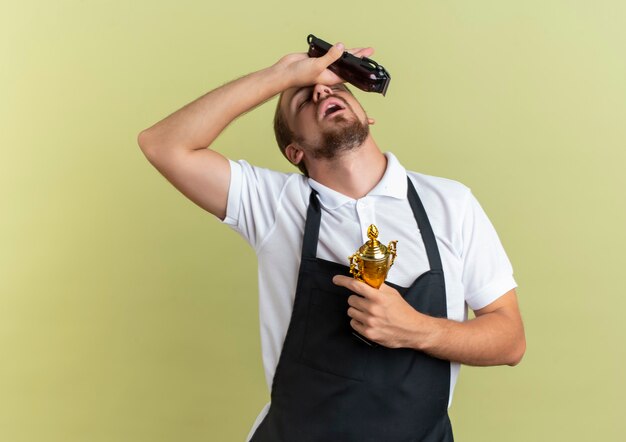 Vermoeide jonge knappe kapper met tondeuses en winnaar beker hand zetten voorhoofd met gesloten ogen geïsoleerd op olijfgroene muur