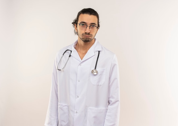 Vermoeide jonge mannelijke arts met optische bril die wit gewaad met stethoscoop op geïsoleerde witte muur met exemplaarruimte draagt