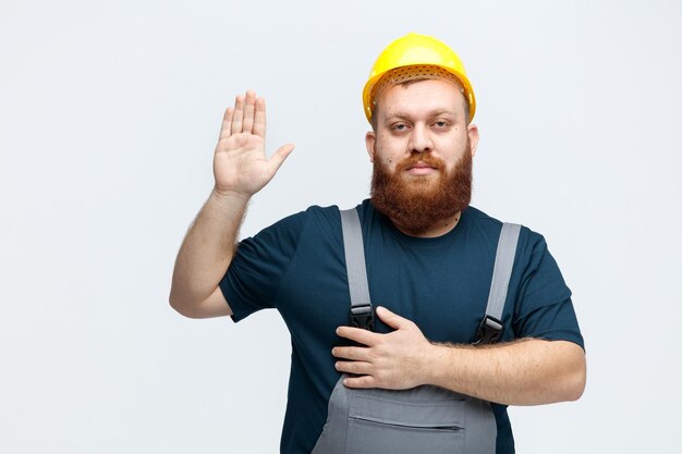 Vermoeide jonge mannelijke bouwvakker met veiligheidshelm en uniform kijkend naar de camera die de hand op de borst houdt en een stopgebaar toont dat op een witte achtergrond wordt geïsoleerd