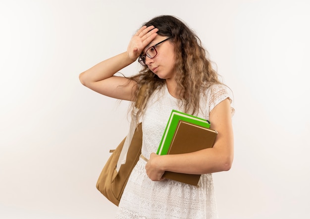 Vermoeide jonge mooie schoolmeisje bril en rug tas bedrijf boeken neerkijkt zetten hand op hoofd geïsoleerd op een witte achtergrond met kopie ruimte