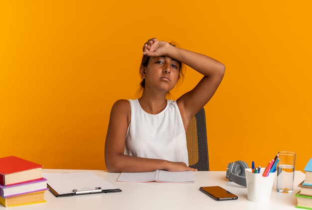 Vermoeide jonge schoolmeisjeszitting bij bureau met schoolhulpmiddelen die pols op voorhoofd zetten die op oranje muur wordt geïsoleerd