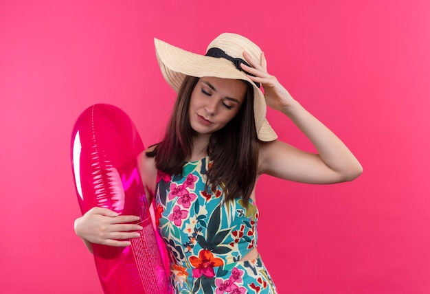 Vermoeide jonge vrouw die hoed draagt die zwemt ring houdt en hand op haar hoofd op geïsoleerde roze muur legt