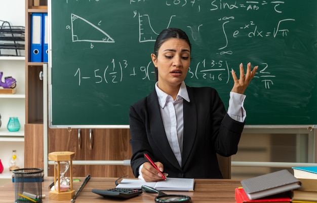 vermoeide jonge vrouwelijke leraar zit aan tafel met schoolbenodigdheden die iets schrijven dat hand in de klas verspreidt