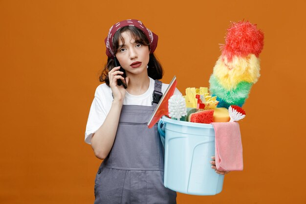 Vermoeide jonge vrouwelijke schoonmaker in uniform en bandana met emmer schoonmaakgereedschap pratend aan de telefoon kijkend naar camera geïsoleerd op oranje achtergrond