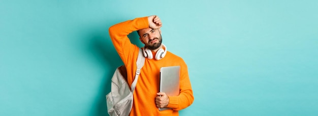 Vermoeide mannelijke student die het zweet van het voorhoofd veegt met laptop en rugzak in oranje trui