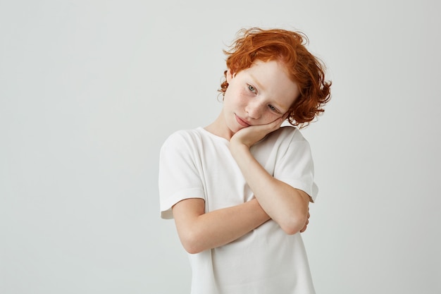 Vermoeide roodharige jongen met sproeten in wit t-shirt op zoek opzij met ontspannen uitdrukking, hoofd met hand houden.