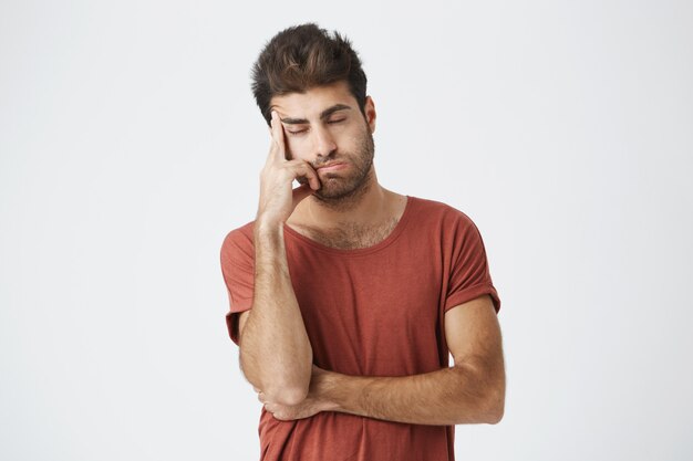 Vermoeide Spaanse gebaarde mens die de rode hand van de t-shirtholding op voorhoofd dragen kijkend zwaar hebbend hoofdpijn na conflictsituatie op het werk. Mensen, stress en migraine.