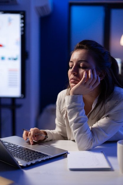 Vermoeide vrouw op afstand die een dutje doet op stoel die op laptop werkt en financiële statistieken analyseert