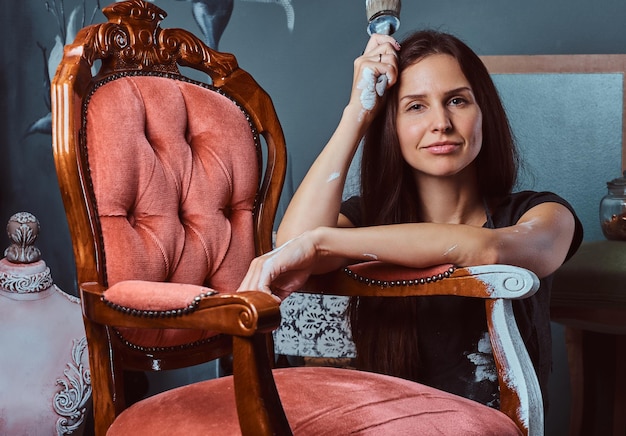 Vermoeide vrouwelijke artiest in schort houdt penseel vast en leunt op een vintage stoel in werkplaats.