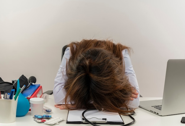 Vermoeide vrouwelijke arts van middelbare leeftijd dragen medische gewaad en stethoscoop zit aan bureau met medische hulpmiddelen Klembord en laptop hoofd zetten bureau geïsoleerd