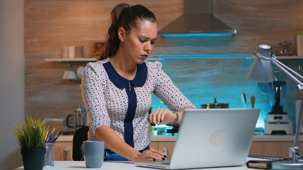 Vermoeide zakenvrouw die overwerkt en thuis koffie drinkt in de moderne keuken. Drukke, gefocuste werknemer die moderne technologie gebruikt, draadloos overwerkt voor werklezen, schrijven, zoeken.