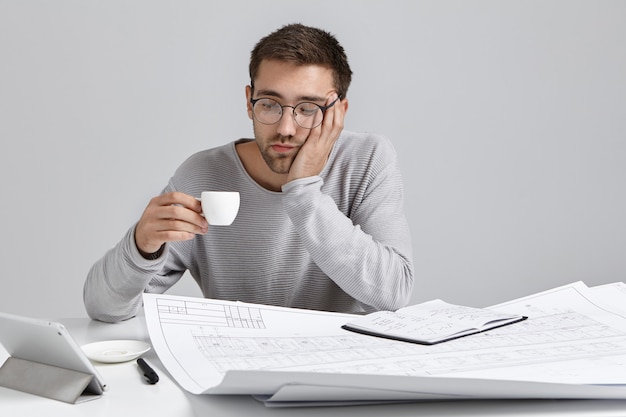 Vermoeidheidsman kijkt de hele avond moe na het werk naar tekeningen, kijkt naar kopje espresso of cappucino