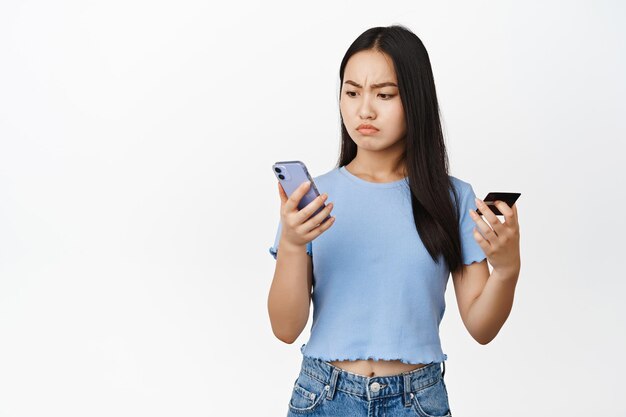 Verontrust Aziatisch meisje met creditcard en smartphone die gefrustreerd kijkt naar haar telefoon en problemen heeft met online betaling op witte achtergrond