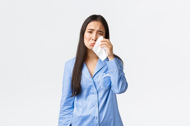 Verontrust dwaas Aziatisch meisje in blauwe pyjama met een gebroken hart, in bed blijven in een slecht humeur, tranen afvegen met tissue, snikken en huilen depressief, rouwend over een witte achtergrond.