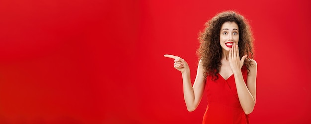 Gratis foto verontruste schattige en tedere dwaze vrouw met krullend kapsel in rode stijlvolle avondjurk die oeps zegt