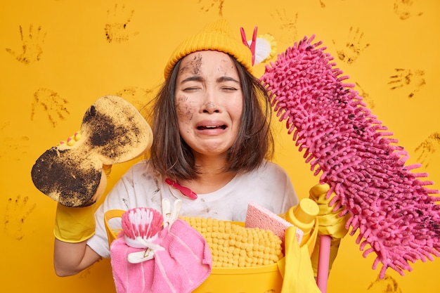 Verontruste vuile vrouw voelt zich overwerkt na de hele dag bezig te zijn met het schoonmaken van poses met spons en spons drukt negatieve emoties uit tegen gele muur