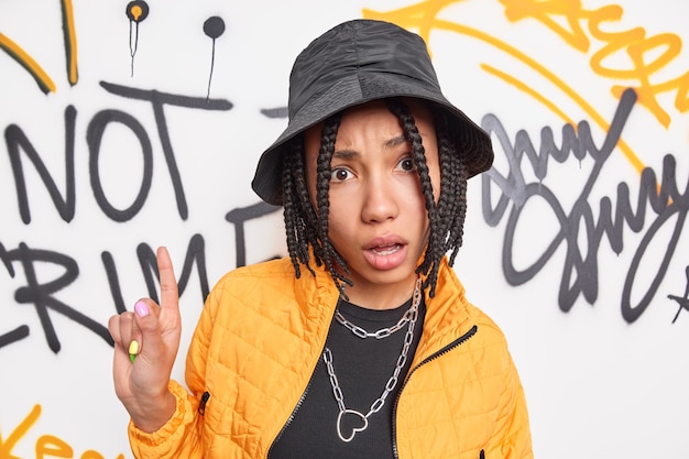 Verontwaardigd hipster meisje met dreadlocks wijst wijsvinger boven draagt zwarte hoed gele jas vormt tegen kleurrijke graffitimuur brengt vrije tijd door in de stad