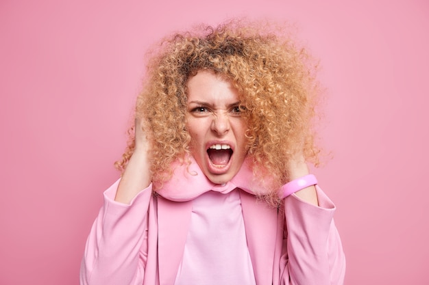 Verontwaardigde gekke jonge vrouw met krullend haar schreeuwt luid omdat ze erg boos is, houdt de mond open en draagt een reiskussen om de nek formele kleding geïsoleerd over roze muur. Menselijke emoties concept emotions