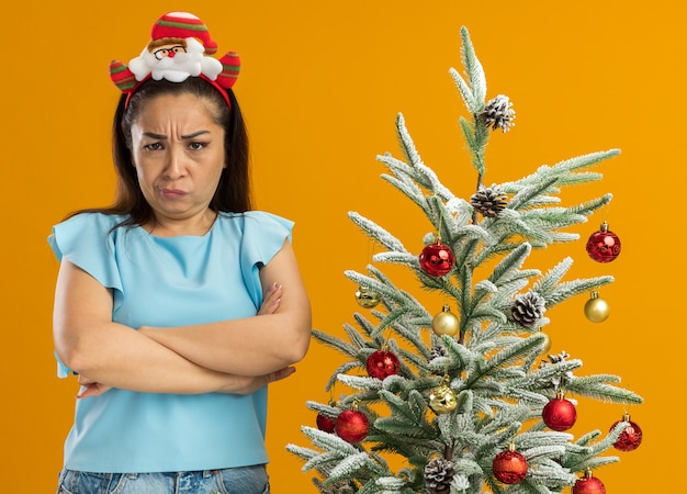 verontwaardigde jonge vrouw in blauwe top met grappige kerstrand op het hoofd camera kijken met fronsend gezicht met gekruiste armen staande naast een kerstboom op oranje achtergrond