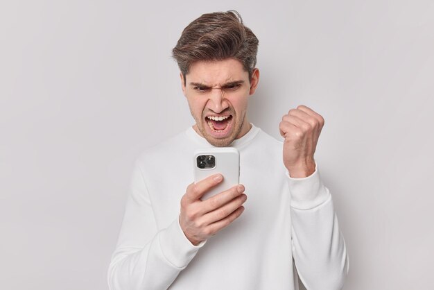 Verontwaardigde man kijkt boos naar smartphone krijgt vervelend bericht balt vuist en roept luid draagt casual jumper geïsoleerd op witte achtergrond ruzie met telefoon drukt negatieve emoties uit