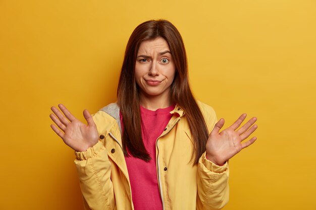 Verontwaardigde onwetende vrouw steekt handpalmen op, poseert onverschillig, heeft een aarzelende uitdrukking in verwarring gebracht, ontevreden over iets, gekleed in een geel jasje, poseert binnen. Menselijke emoties en reactieconcept