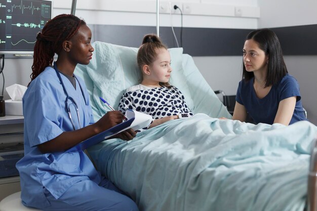 Verpleegkundige in de kinderkliniek die aantekeningen maakt over succesvol herstel in de patiëntenkamer. Moeder die attent is op de verpleegster die spreekt over de gezondheidsrisico's van kleine meisjes terwijl ze op de herstelafdeling ligt.