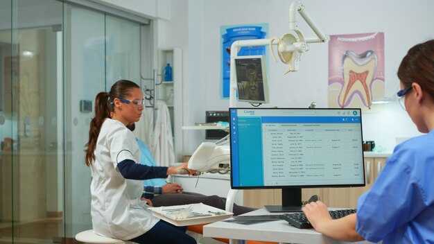 Verpleegkundige typt op de computer, maakt afspraken terwijl de tandarts vóór het onderzoek de patiënt in de stomatologiestoel praat. Tandarts en verpleegster werken samen in moderne stomatologische kliniek