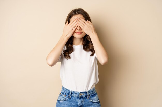 Verrassing. Gelukkige jonge vrouw sloot ogen met handen en wacht op cadeau, staande over beige achtergrond