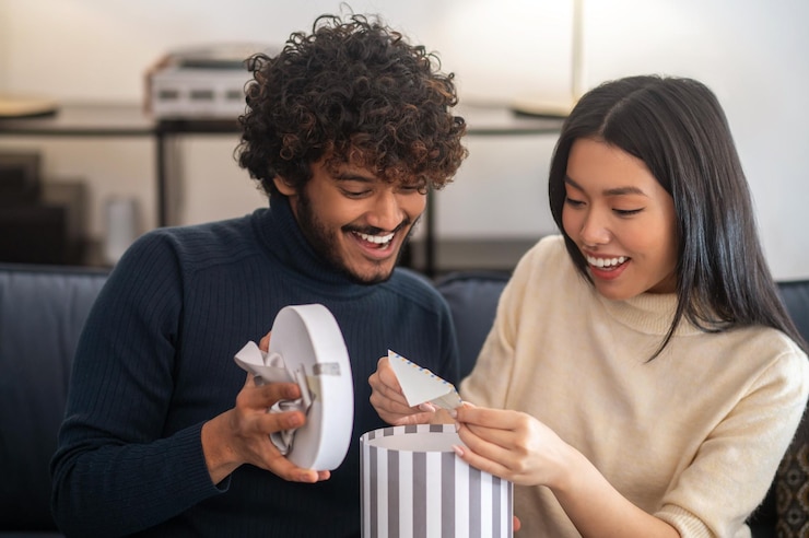 Gratis Foto verrassing, vreugde. glimlachende attente indiase man met krullend haar die de lichtbak van een geschenk opent en aangenaam verraste mooie aziatische vrouw die een brief terugtrekt die op de bank zit