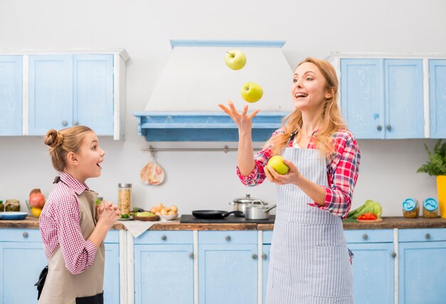 Verrassingsmeisje die haar moeder bekijken die de groene appel in lucht bij keuken werpen