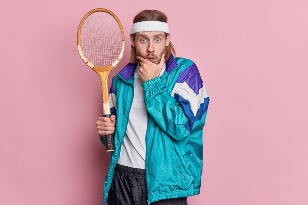 Verrast actieve tennisser houdt racket vast kijkt verbaasd houdt kin vast heeft dikke haren draagt hoofdband en sportkleding.