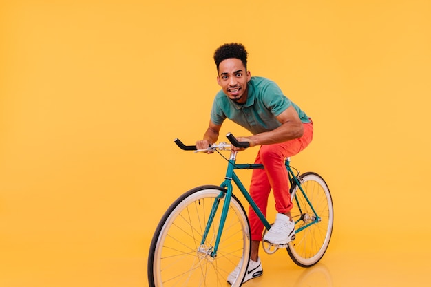 Verrast Afrikaanse man in rode broek rijden op de fiets. Indoor foto van grappige zwarte jonge man zittend op de fiets.