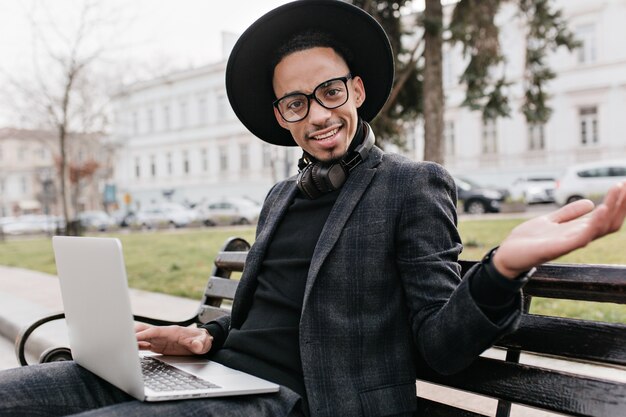 Verrast Afrikaanse student zittend op een bankje met computer. Buiten foto van grappige zwarte mannelijke freelancer met behulp van laptop voor werk.