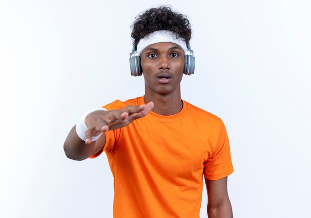 Verrast, afro-amerikaanse sportieve man met hoofdband en polsbandje en koptelefoon die hand naar camera reikt