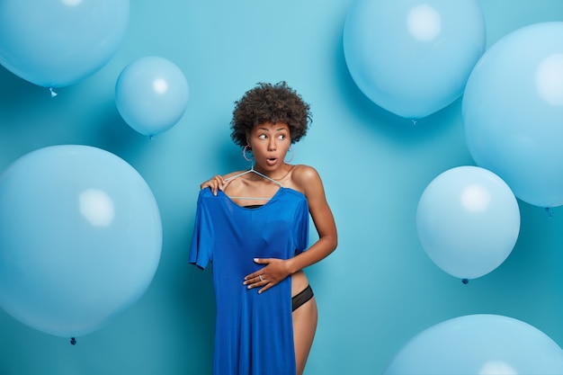Verrast Afro-Amerikaanse vrouw kiest jurk uit haar garderobe, kiest outfit om te dragen bij speciale gelegenheden, staat uitgekleed en verbergt halfnaakt lichaam als iemand komt, geïsoleerd over blauwe muur