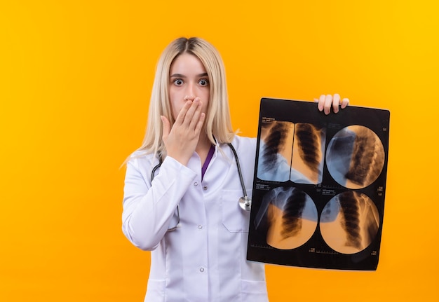 Gratis foto verrast artsen jong meisje die stethoscoop in medische toga dragen die x-ray en behandelde mond met hand op geïsoleerde gele achtergrond houden