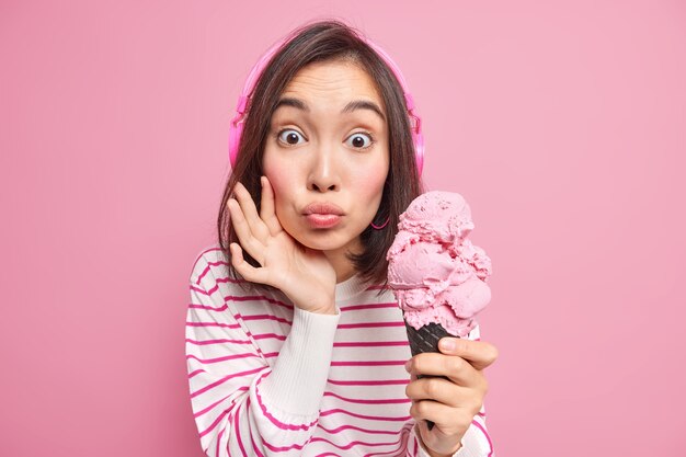 Verrast Aziatische vrouw houdt lippen afgerond eet heerlijk ijs wil koud bevroren dessert eet draagt koptelefoon op oren casual gestreepte trui geïsoleerd over roze muur. Zomer tijd.