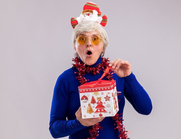 Verrast bejaarde vrouw in zonnebril met santa hoofdband en slinger rond nek opening kerstcadeau doos geïsoleerd op een witte muur met kopie ruimte