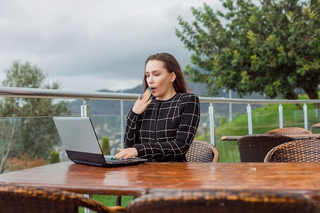 Verrast bloggermeisje kijkt naar haar laptopscherm door haar hand op de mond te houden op de achtergrond van de natuurweergave