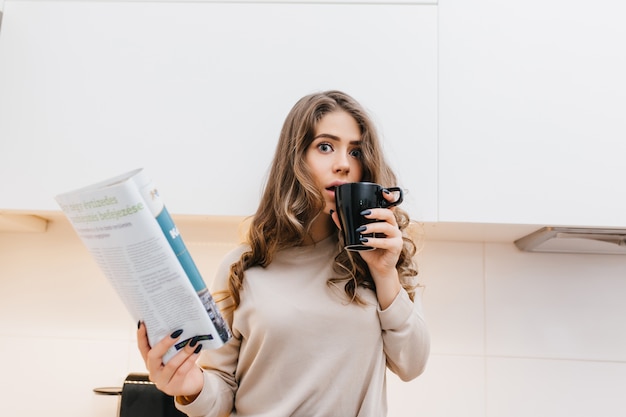 Verrast brunette vrouw koffie drinken tijdens het lezen van tijdschrift in de ochtend