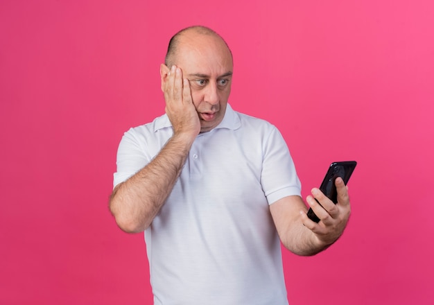 Verrast casual volwassen zakenman houden en kijken naar mobiele telefoon en hand houden op gezicht geïsoleerd op roze achtergrond met kopie ruimte