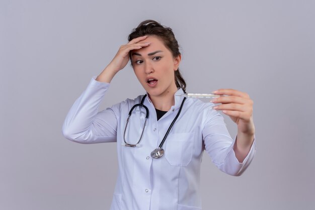 Verrast dokter jong meisje medische jurk dragen stethoscoop houden thermometer en legde haar hand op voorhoofd op witte achtergrond
