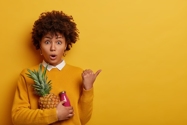 Verrast emotionele vrouw met Afro haar houdt verse ananas en fles roze smoothie