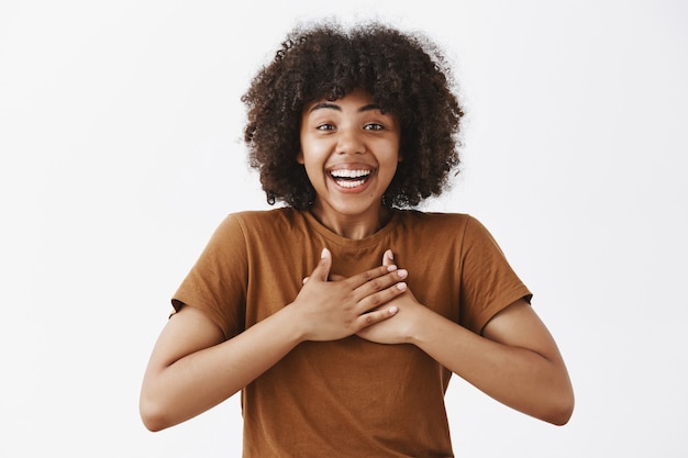 Verrast en dankbaar aantrekkelijke tedere afro-amerikaanse vrouw in trendy bruin t-shirt hand in hand op hart en breed glimlachend van verbazing en vreugde ontvangen onderscheiding over grijze muur