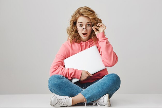Verrast en geschokt meisje opstijgen bril, zittend met laptop op de vloer
