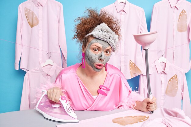 Verrast gekrulde huisvrouw ondergaat schoonheidsbehandelingen terwijl ze huishoudelijk werk doet, past een kleimasker op het gezicht vast, houdt een plunjer, strijkijzers, kleren of was op de strijkplank, draagt een slaapmasker en een kamerjas