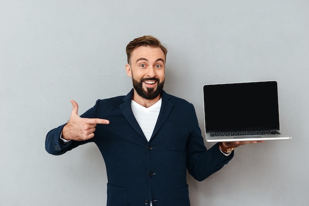 Verrast gelukkig bebaarde man in zakelijke kleding met lege laptop computerscherm en wijzend op hem over grijs