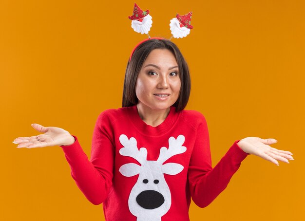 Verrast jong Aziatisch meisje dat de hoepel van het Kerstmishaar met sweater spreidende handen draagt die op oranje muur worden geïsoleerd